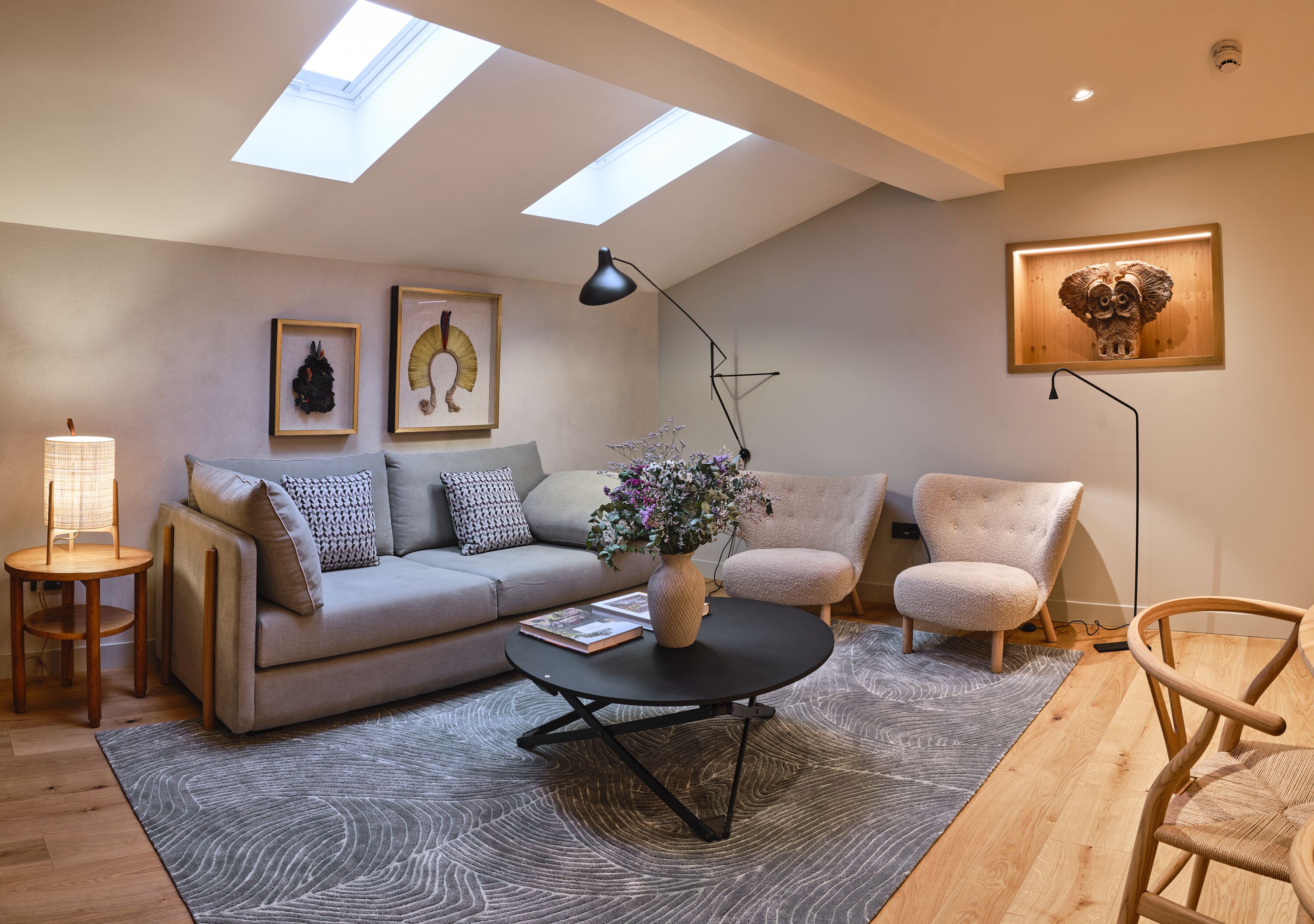 A modern living room with a sofa, armchairs, a coffee table, and a floor lamp. There are skylights on the ceiling, a rug, and a framed picture on the wall.
