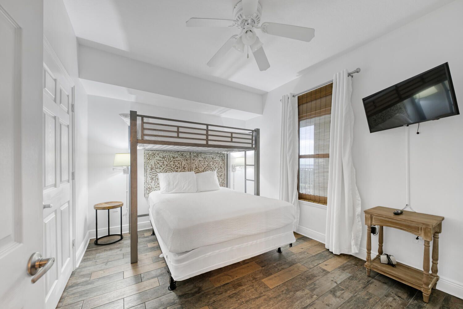 A bedroom with a bunk bed, a television mounted on the wall, a wooden floor, and a window with curtains.