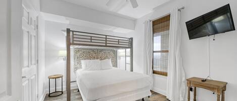 A bedroom with a bunk bed, a television mounted on the wall, a wooden floor, and a window with curtains.