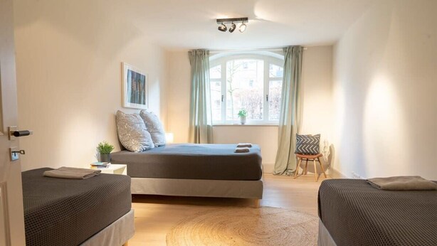 Une chambre avec deux lits simples, une grande fenêtre avec des rideaux, une table de chevet agrémentée d’une plante et un tapis rond en fibres tressées.