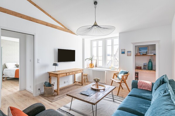 A modern living room with a wooden ceiling, a large window, a TV mounted on the wall, a sofa, a coffee table, and a desk with a chair. - Sieck - Joli Appartement Vue mer (Carantec)