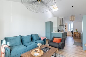 A modern living room with a blue sofa, a wooden coffee table, and a grey armchair. There is a kitchen area with a dining table and chairs visible in the background.