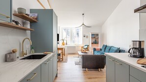 Una cocina moderna con horno integrado, un lavamanos y estantes de madera. Hay un comedor con mesa y sillas, y un sofá en la sala de estar.