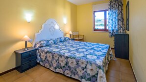 Une chambre à coucher avec une couvre-lit à motifs floraux, une table de chevet avec une lampe, une commode et une fenêtre avec des rideaux.