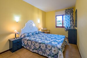 A bedroom with a floral bedspread, a nightstand with a lamp, a dresser, and a window with curtains.
