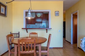 A dining area with a round wooden table and chairs, a hanging light fixture, a wall clock, and a window with a wooden frame.
