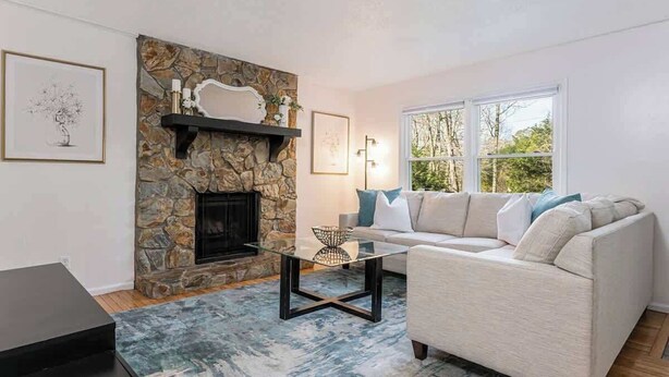 A living room with a stone fireplace, a white sofa with blue cushions, a glass coffee table, and a large window. - 4BR Home Minutes to DOWNTOWN, RAD & BILTMORE (Arden)