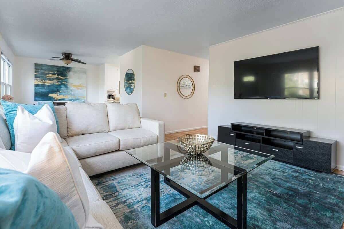 Una sala de estar moderna con un sofá seccional, una mesa de centro de vidrio, un televisor de pantalla plana y un ventilador de techo.