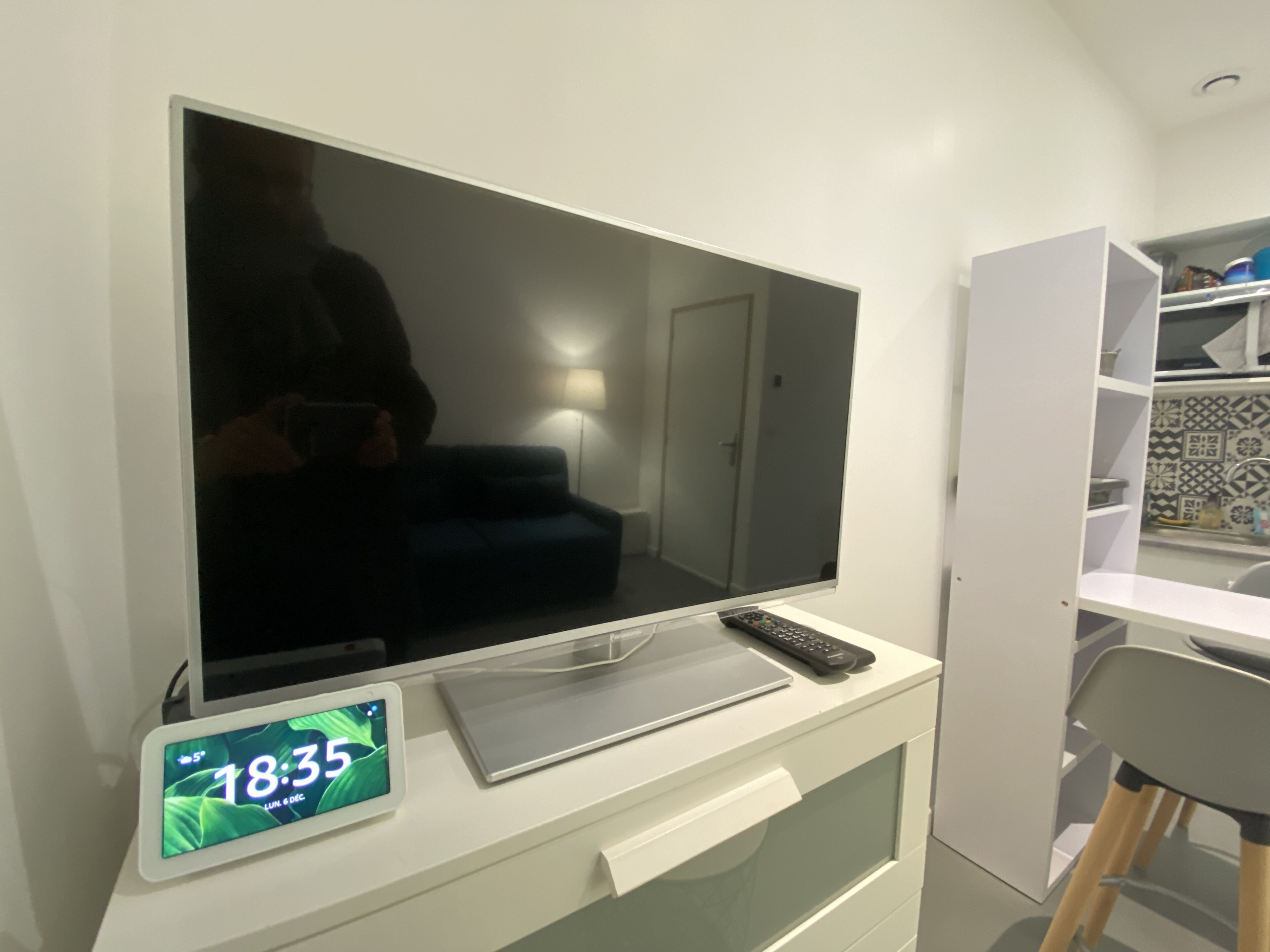 A modern room with a large flat-screen TV mounted on a white wall, a digital clock displaying 18:35, and a remote control on the TV stand.