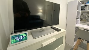 A modern room with a large flat-screen TV mounted on a white wall, a digital clock displaying 18:35, and a remote control on the TV stand. - #Casamara 1 2-bed apartment | Netflix (Amiens)