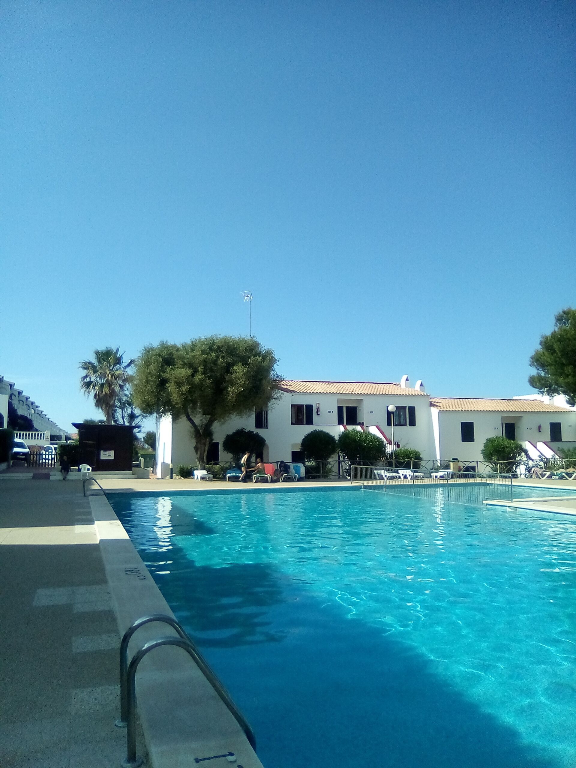 Une piscine à l’eau d’un bleu cristallin, entourée de bâtiments blancs aux toits recouverts de tuiles rouges, et de quelques arbres.