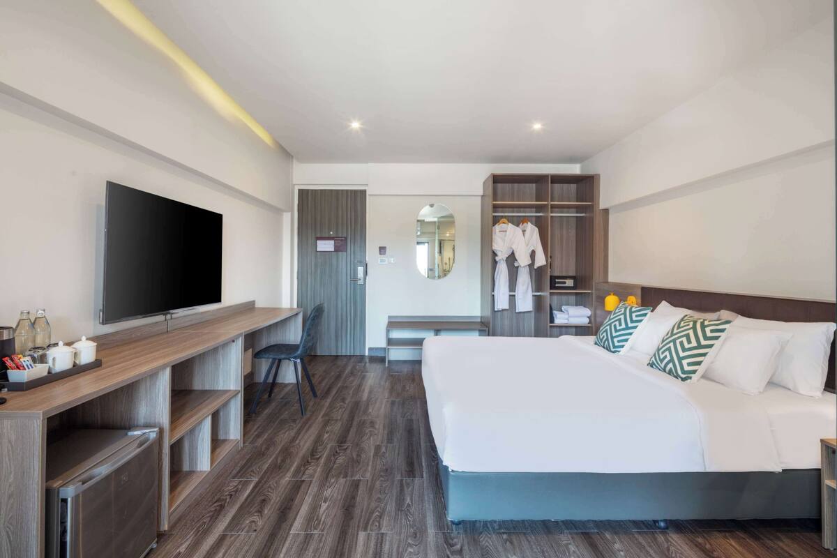 A modern hotel room with a large bed, a TV, a desk with a chair, and a wardrobe with hanging robes.