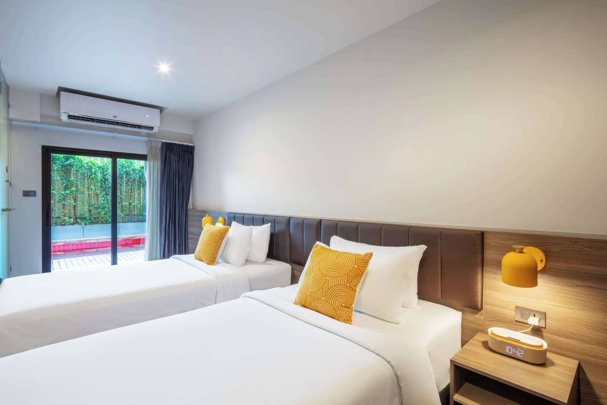 A hotel room with two beds, a bedside table with a lamp, and a view of greenery outside.