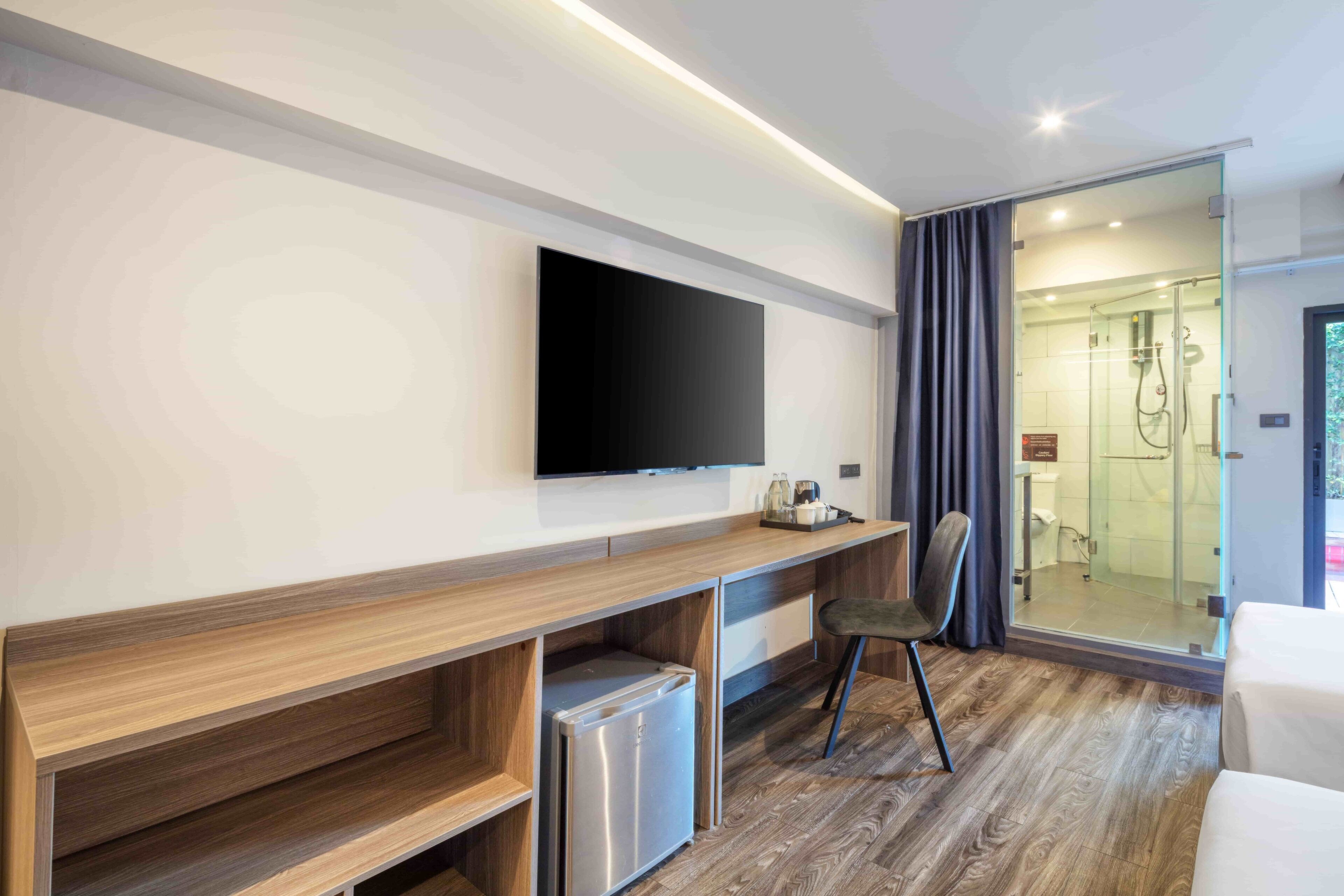 A modern hotel room with a wooden desk, a flat-screen TV, a mini-fridge, a chair, and a glass shower area.