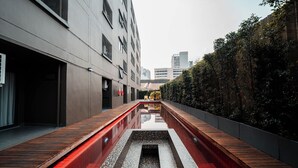Een smalle steeg met een reflecterend waterpartij, houten bankjes en een haag aan één kant.