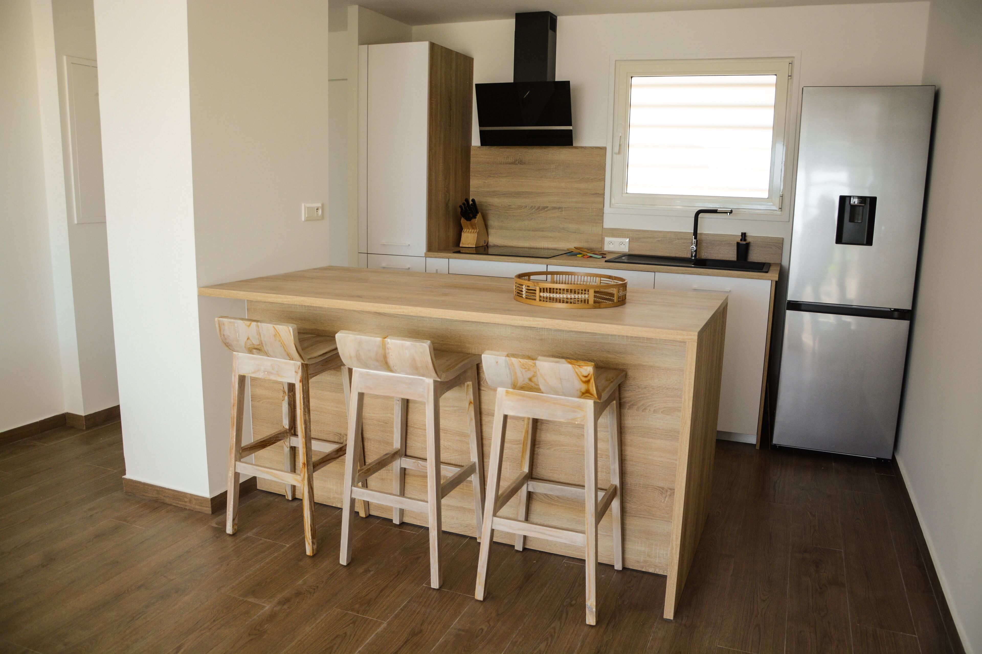 A modern kitchen with a central island, bar stools, a stainless steel refrigerator, and a window above the sink.