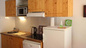 Cocina compacta con armarios de madera, microondas, hervidor, tostadora y un refrigerador pequeño.