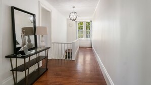 A hallway with a wooden floor, a black console table with a lamp, a mirror, and a window with a view of greenery. - NEW! Historic Townhome - Live among the Mansions 3BR (Newport)