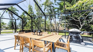 Relax in the serene patio area surrounded by lush greenery and palm trees, complete with a barbecue grill and pergola for shade.