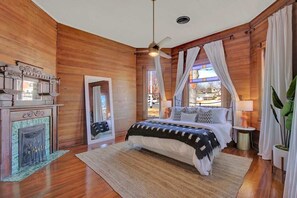 A bedroom with a wooden floor, a bed with a black and white patterned bedspread, a ceiling fan, and a fireplace with a decorative mantle.