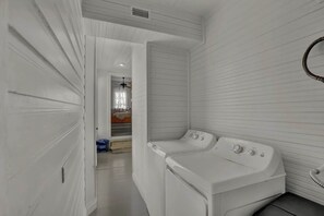 A laundry room with two white washers and dryers, a vent on the ceiling, and a door leading to another room.