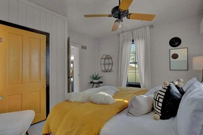 Une chambre à coucher avec un lit, un ventilateur de plafond, une fenêtre avec des rideaux, une porte et une petite table sur laquelle est posée une plante.