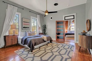 Ein Schlafzimmer mit Bett, Deckenventilator, großem Fenster mit Vorhängen, Nachttisch und Beistelltisch.