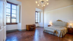 Ein geräumiges Zimmer mit einem großen Bett, einem Holztisch und einem Stuhl. Zwei Fenster sorgen für natürliches Licht, und eine Kristallleuchter hängt von der Decke.