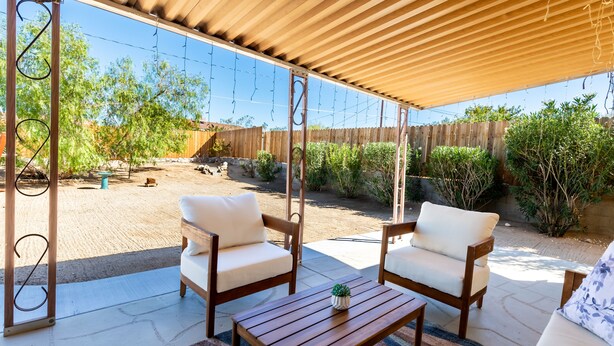 A covered patio with two wooden chairs and a table, a wooden swing set, and a view of a backyard with a wooden fence and greenery. - Eternity's Edge -Hike/Stargaze, Peaceful, Polished (Twentynine Palms)