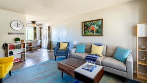 A living room with a sofa, coffee table, and a bookshelf. There is a dining area with a table and chairs in the background. - Eternity's Edge -Hike/Stargaze, Peaceful, Polished (Twentynine Palms)