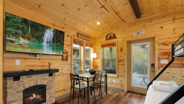 Interior de una cabaña de madera con chimenea, una mesa de comedor con sillas, un televisor mostrando una cascada y un cartel que dice 'WELCOME'.