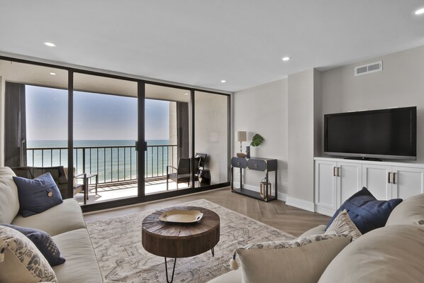 Relax in a spacious living area with a sofa, armchairs, and flat-screen TV, leading to a balcony with a sea view.