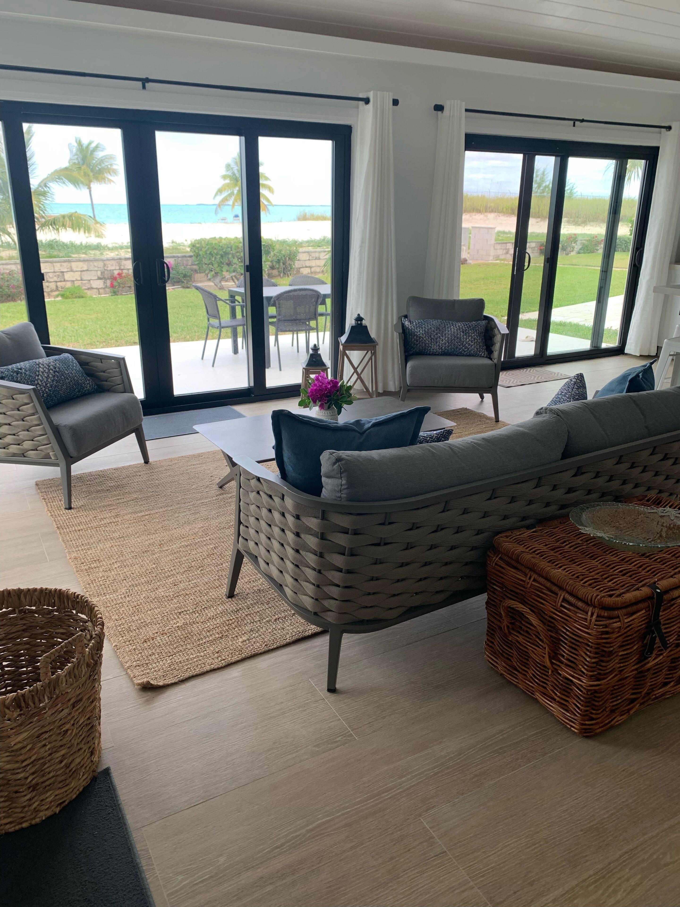 A spacious living room with a large sliding glass door leading to a balcony, wicker furniture, and a view of the beach.
