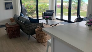 Ein modernes Wohnzimmer mit einer Couch, Sesseln und einem Esstisch. Große Schiebeglasfenster führen in einen Garten mit Blick auf den Ozean.
