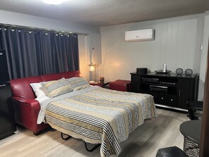 Habitación de hotel con sofá de cuero rojo, cama con ropa de cama a rayas, una mesita con lámpara, mueble de entretenimiento negro y aire acondicionado instalado en el techo.