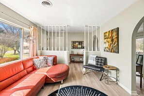 A modern living room with a red sofa, a black leather chair, and a wooden coffee table. There is a large window with a view of trees and a road.