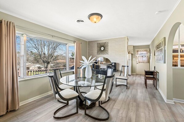 A dining area with a glass table and chairs, a kitchen in the background, and a view of the outdoors.