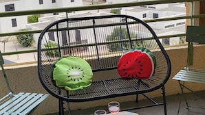 Un balcon avec une chaise et une table en métal, deux coussins et deux verres de vin. On peut admirer, depuis le balcon, une vue sur une plage et des bâtiments.