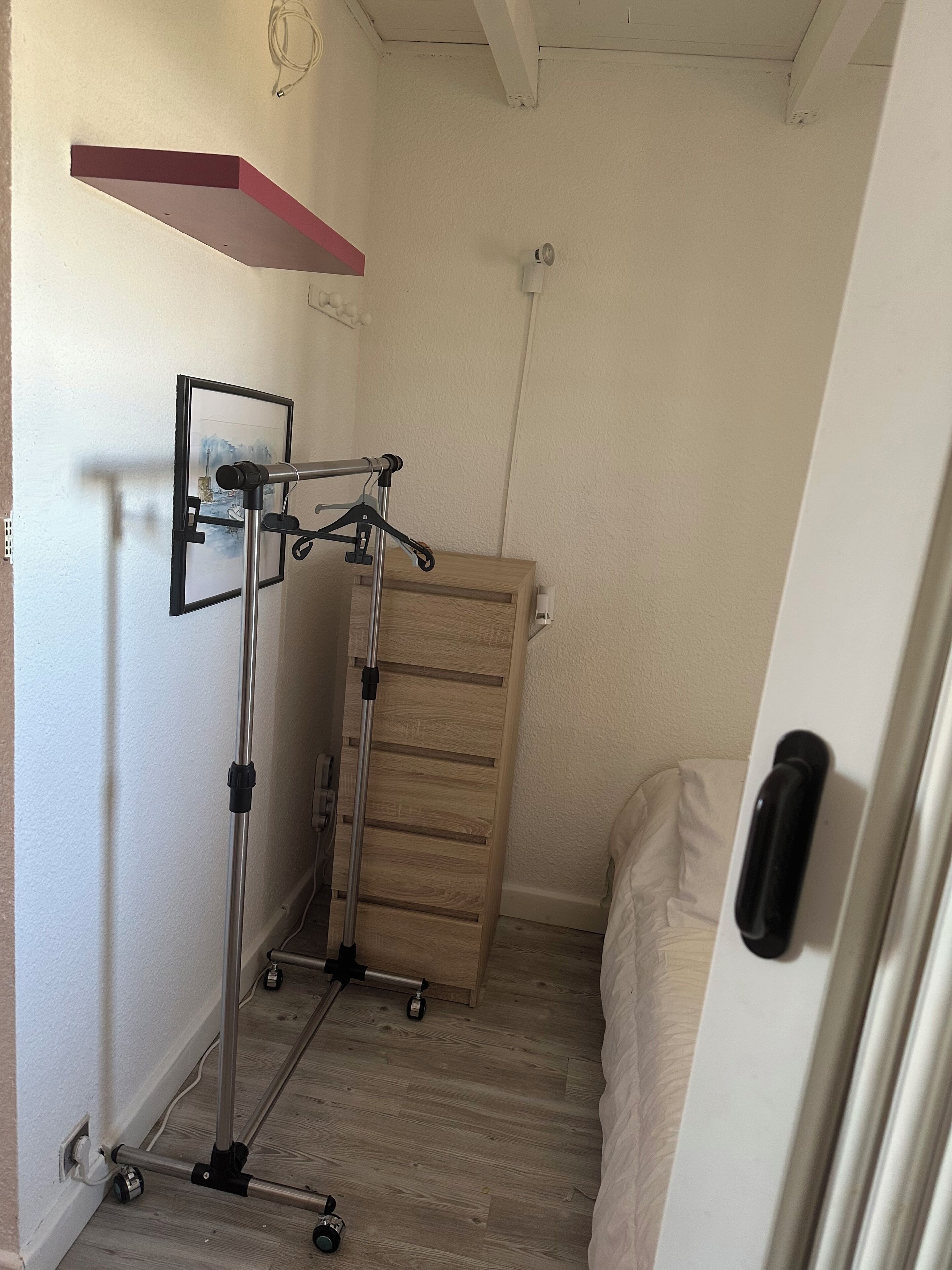Une chambre à coucher comprenant un lit, une commode en bois, une étagère fixée au mur et un trépied.