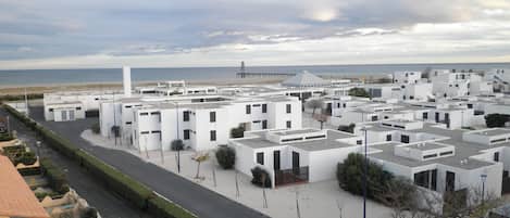 A coastal residential area with modern white buildings, a beach in the background, and a clear sky.