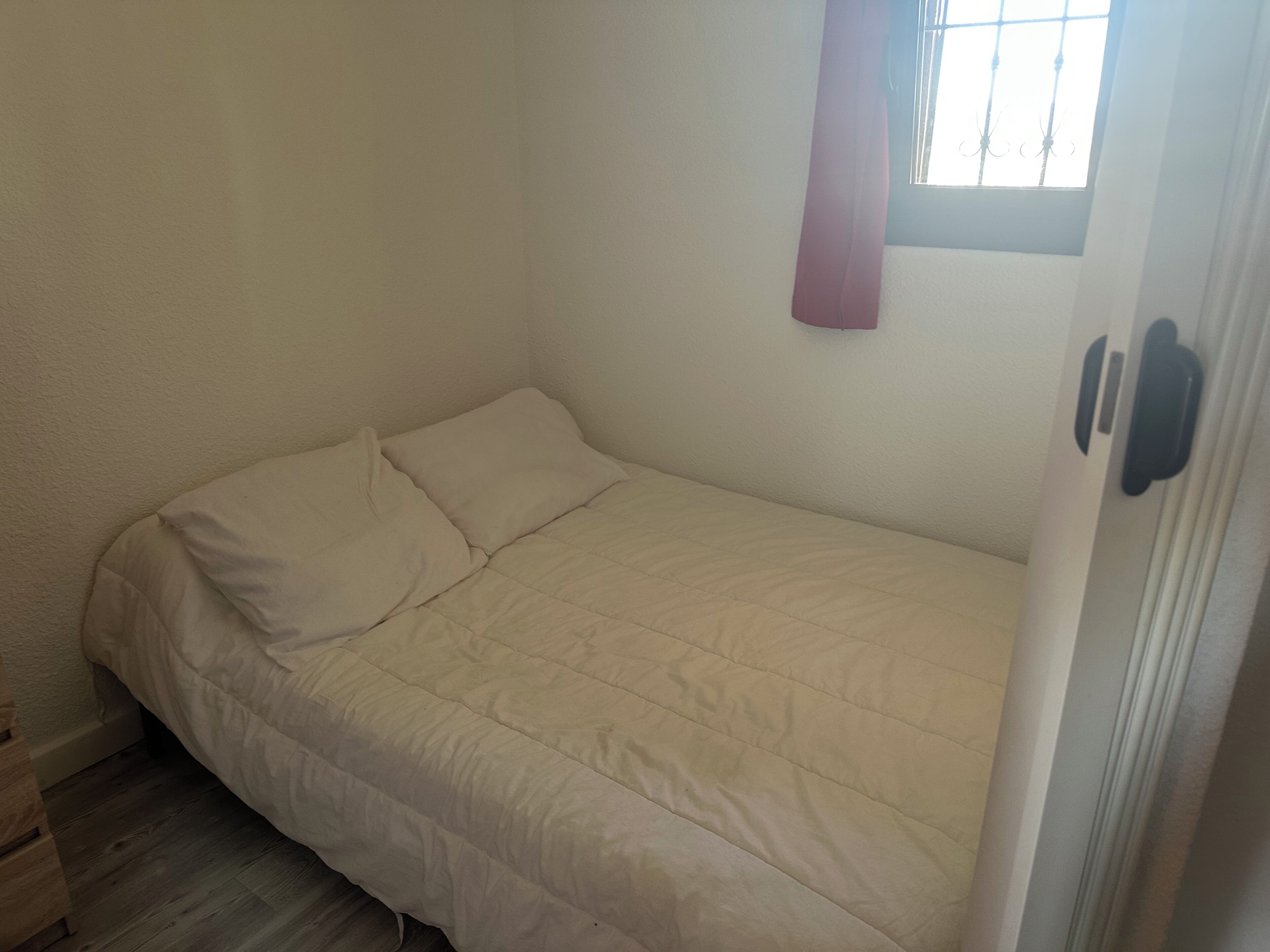 Una habitación pequeña con una cama individual, pared blanca sencilla, ventana con rejas y suelo de madera.