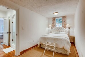 Un dormitorio con cama blanca, mesita de noche, lámpara y ventana con cortinas.