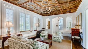 A spacious living room with a high wooden ceiling, large windows with blinds, floral-patterned sofas, and a chandelier.