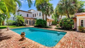 A pool area with a clear blue pool, surrounded by a brick patio and lush greenery, including palm trees.