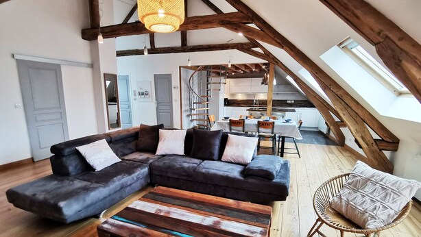 A loft-style living area with a large sectional sofa, a wooden coffee table, and a wicker chair. A kitchen is visible in the background with a dining table and chairs. - Ô calme - 100m² apartment 15 min from Orléans (Olivet)