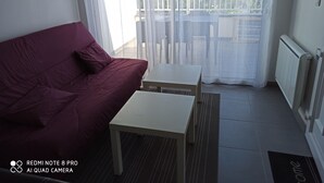 Una habitación con un sofá rojo, una mesita blanca y vistas a través de una ventana con cortina blanca.