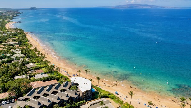 Vue aérienne d’un complexe hôtelier en bord de mer, avec de nombreux bâtiments, une eau turquoise et limpide, et une plage de sable.