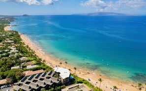 Vue aérienne d’un complexe hôtelier en bord de mer, avec de nombreux bâtiments, une eau turquoise et limpide, et une plage de sable.