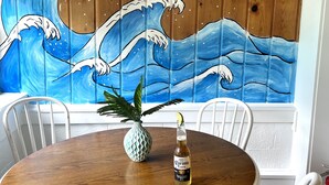 A wooden table with a bottle of Corona Extra beer, a pineapple, and a white vase. A mural of a wave is on the wall behind the table.