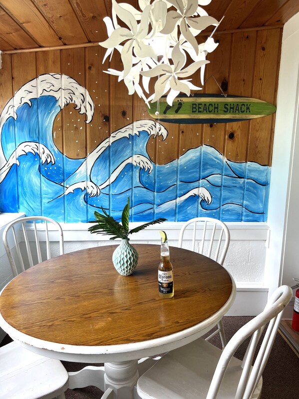Una mesa de madera con una botella de cerveza Corona Extra, una piña y un jarrón blanco. Un mural de una ola adorna la pared detrás de la mesa.